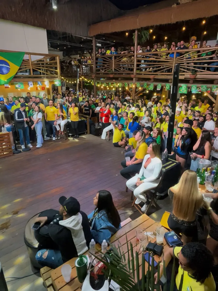 Casa cheia no deck de dois andares assistindo ao jogo, com bandeira do Brasil e luzes penduradas