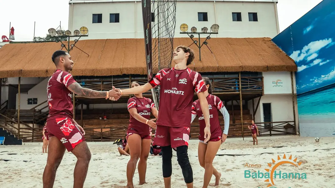 Quadra de areia da arena com atletas jogando diante do painel de praia